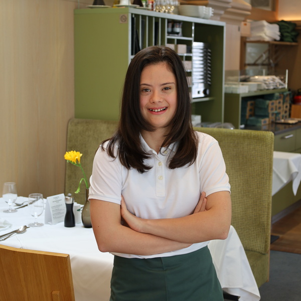Young woman with long dark hair standing in a restaurant, smiling and with her arms crossed.