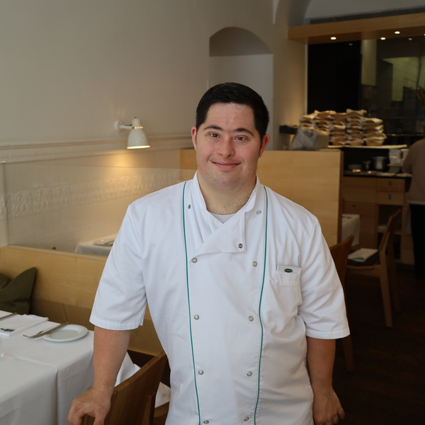 A chef in a white uniform stands smiling in a modern, well-lit dining area of a restaurant.