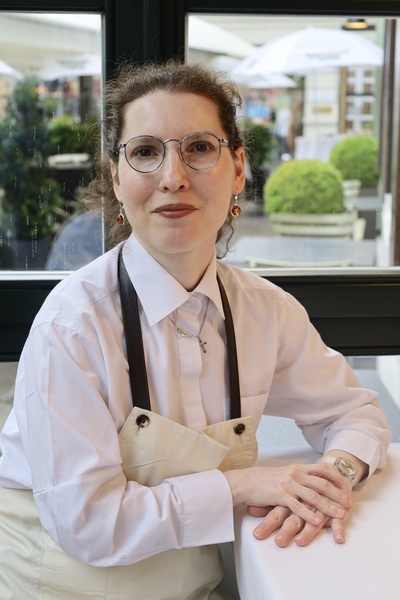 Woman with glasses and apron sits at a table in a bright café, hands folded, smiling gently into the camera.