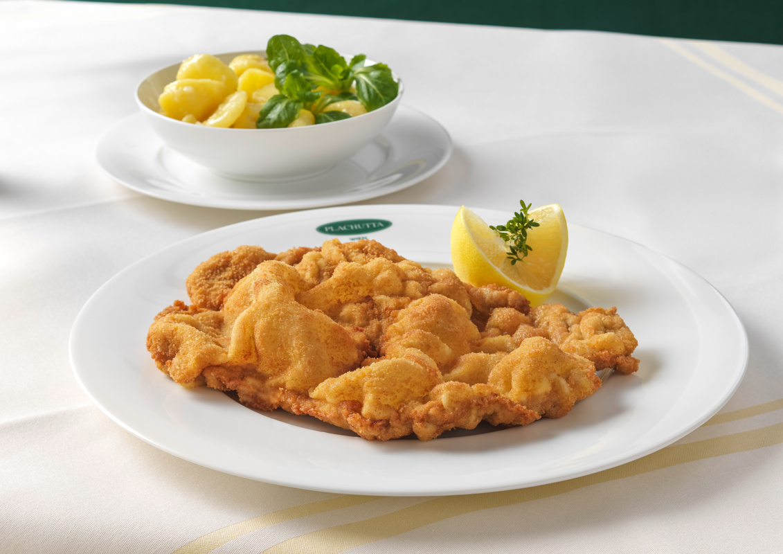 Ein Teller paniertes Schnitzel mit Zitronenspalte, serviert mit Kartoffeln und Grünzeug.